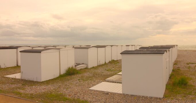 Pan shot across rows of white wooden beach huts on pebble shore in Le Havre, Normandy. Moody overcast sky, muted tones.