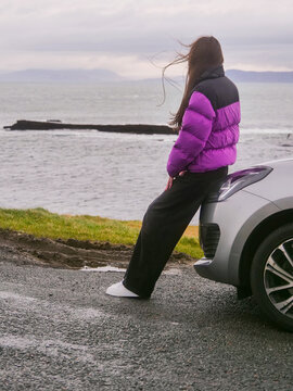 Young tourist woman in purple warm jacket is standing by silver rented car and looking at stunning nature scenery with ocean and mountains in the background. Travel industry. Mullaghmore, Ireland.