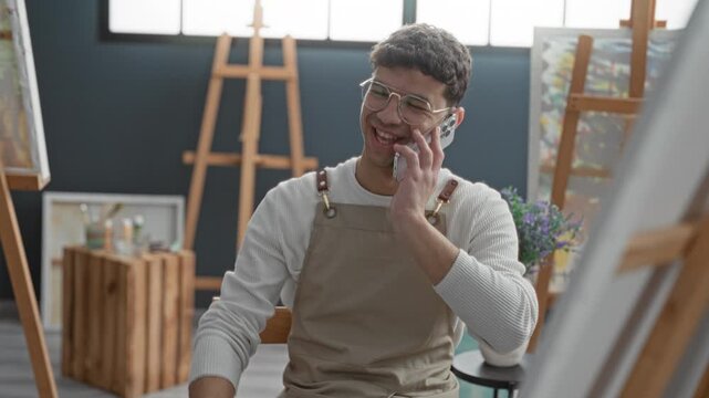 Man in apron smiling, holding smartphone to ear and gesturing with left hand amid easels, canvases, brushes and crate in an art studio; joyful conversation.