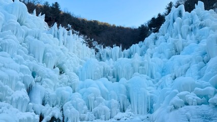Stunning ice wall with giant icicles and frozen formations in a winter valley under a clear blue sky. © Thararut