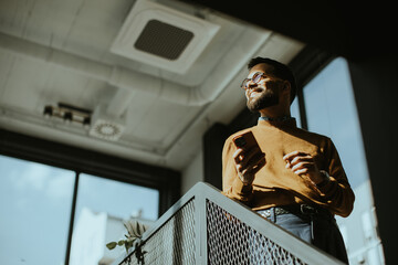 Man stands on a balcony looking out at the city while holding a smartphone during daytime © BGStock72
