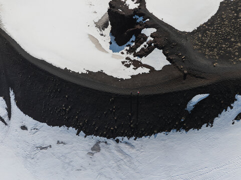 Aerial view of the volcanic crater rim on the south side of Mount Etna with black ash, patches of white snow and a hiker standing on the edge in Nicolosi, Sicilia, Italy.