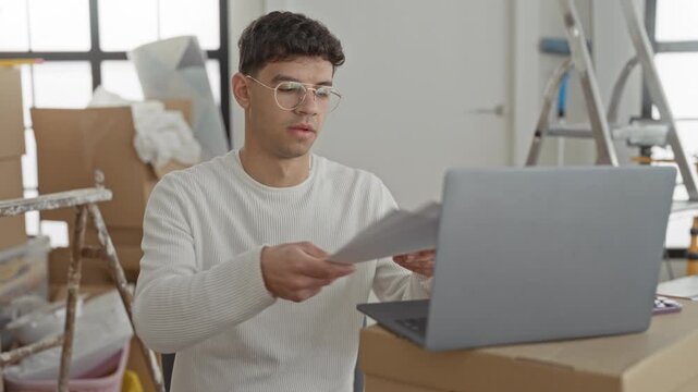 Man at a makeshift desk with laptop and cardboard boxes reading papers then leaning back with hands behind head in a building; relief settling.
