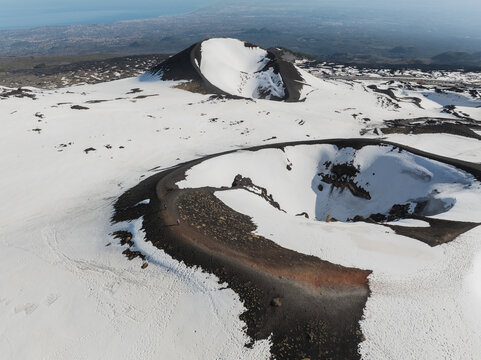 Aerial view of the snow-covered volcanic craters on the south side of Mount Etna with dark volcanic ash and a distant view of the coastline in Nicolosi, Sicilia, Italy.