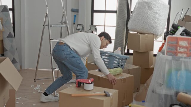 Man clutching lower back while bending over packed cardboard boxes and a ladder amid moving supplies inside a building; pain backache strain.