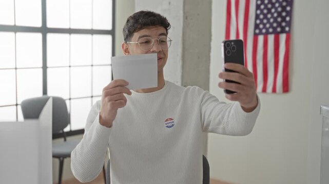 Man taking selfie while holding smartphone and ballot inside a voting building, showing voted sticker on chest; civic pride.