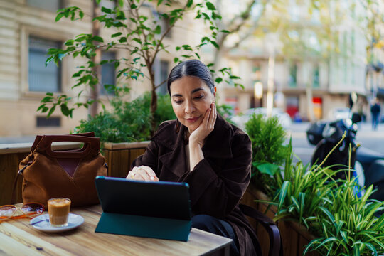 Thoughtful Hispanic woman working outdoors at cafe table with tablet, hand on face, reflecting focus, mindfulness, and urban freelance lifestyle surrounded by greenery.