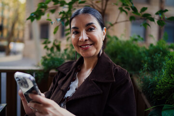 Confident Hispanic woman smiling at camera while holding smartphone in both hands, showing joy, approachability, and digital connection during mobile freelance workflow. © BullRun