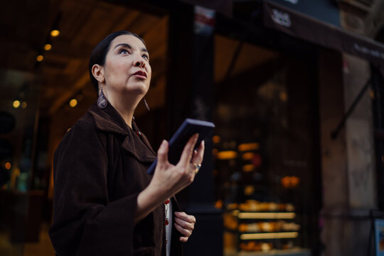 Confident Hispanic woman looking up while recording a voice note, standing near shop window, symbolizing digital focus and clarity in modern communication habits.