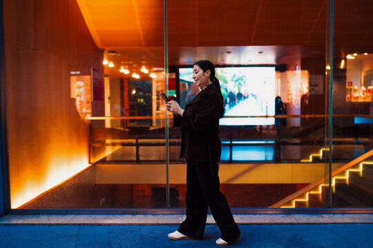 Hispanic woman walks past glass wall while focused on smartphone, representing on-the-go connectivity, tech fluency and urban independence in mobile professional life.