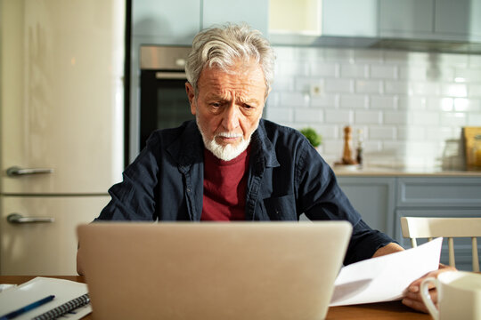 Worried senior man reviewing bills on laptop at home kitchen