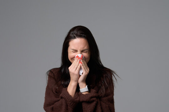 Sick woman sneezing into white tissue holding napkin with both hands isolated on grey background
