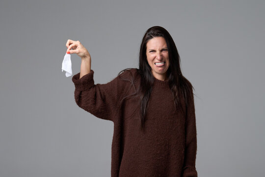 Woman holding used tissue at distance with look of disgust and aversion on grey background