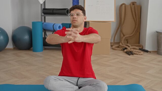 Man stretching hands forward while seated crosslegged on yoga mat in studio with exercise balls and ropes visible; calm focus.