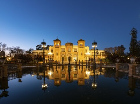 Illuminated art museum Museo de Artes y Costumbres Populares de Sevilla reflected in a fountain, blue hour, Plaza de America, Seville, Andalusia, Spain