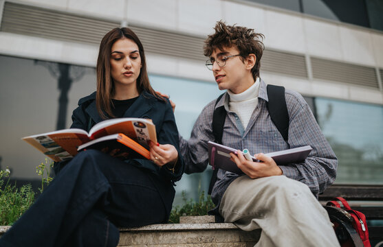 Two high school students sit on a bench and study from books and notebooks. A student tutors and discusses notes with a classmate in an outdoor campus setting.