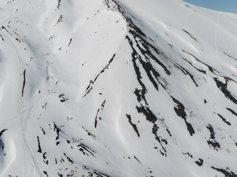 Aerial view of the snow-covered slopes of Mount Etna south side with ski lift pylons and dark volcanic rock patches in Nicolosi, Sicilia, Italy.