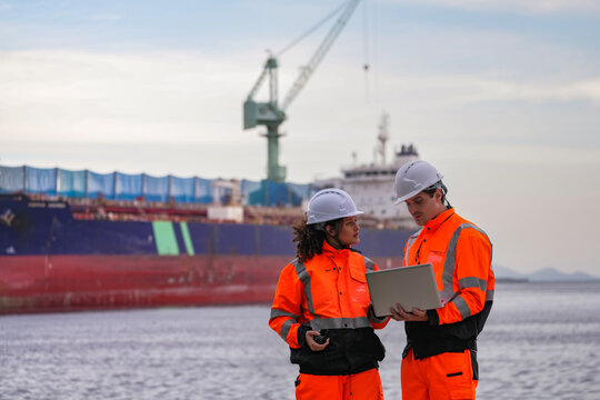 Diverse industrial engineers in orange safety uniforms and hard hats collaborating with a laptop at a shipping port. Happy coworkers discussing logistics near a large cargo ship.