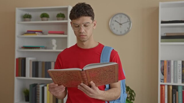 Young man holding book and backpack places finger to lips shushing in building, wearing red shirt and glasses; quiet focus.