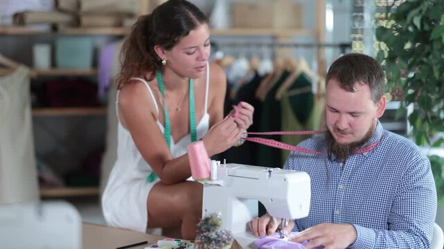 Cheerful young female tailor sitting on work table and playfully teasing male coworker working at sewing machine, making him look shy and slightly confused in workshop
