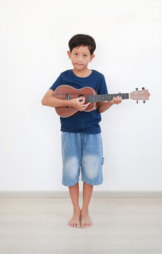 Portrait of happy young Asian boy playing the ukulele, learning a new hobby at home against white wall.
