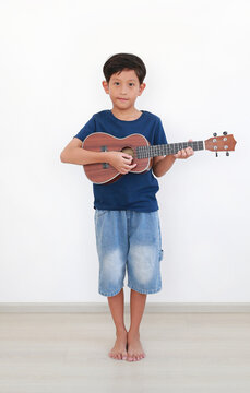 A young Asian boy standing in blue t-shirt and denim shorts, happily playing a brown ukulele against a white wall.