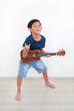 Cheerful young Asian boy in blue t-shirt and denim shorts strikes an energetic pose while holding ukulele and making a rock hand sign against white wall.