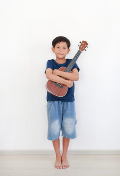 A young boy, barefoot and smiling, stands holding a ukulele closely in his arms against a plain white wall.