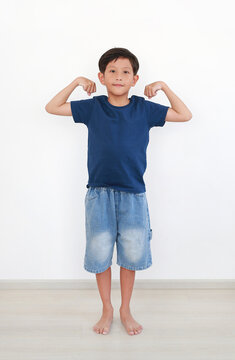 Portrait of young Asian boy in navy blue t-shirt and denim shorts posing with his arms up, showing his muscles on white wall background in the room.