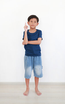 Portrait of young Asian boy in a blue t-shirt and denim shorts stands confidently, pointing upwards with his index finger against white wall background.