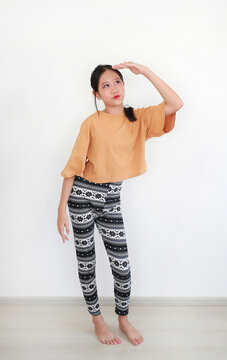 Young Asian girl in stylish orange top and patterned leggings strikes a playful pose, shading her eyes with her hand as if searching for something in distance against white wall.