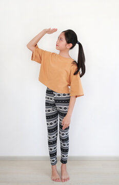 Young girl with ponytail, wearing mustard yellow crop top and patterned leggings, is playfully posing by placing her hand above her eyes as if looking into the distance on white wall.
