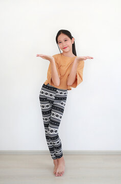 An attractive Asian young girl in stylish outfits is standing barefoot on wooden floor while leaning on white background, showing her hands.