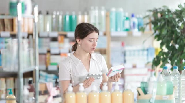 Thoughtful young woman examining tablet blister and packaging at pharmacy counter, carefully checking form, quantity, or expiration date before purchase. High quality 4k footage