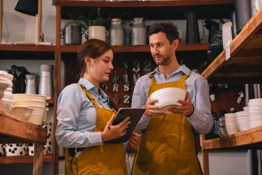 Store inventory review in ceramic shop with two worker holding bowl and tablet, focused retail supply check in warm light