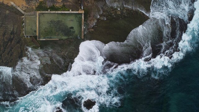 Aerial view of Newport Rock Pool with crashing white waves against dark coastal rocks and swimmers in the rectangular ocean pool in Sydney, New South Wales, Australia.