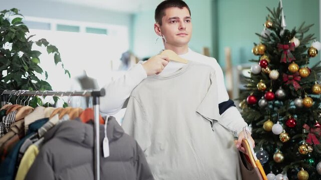 Guy chooses comfortable basic T shirts for every day against the background of the Christmas tree. Buying cotton clothes in the store. High quality 4k footage
