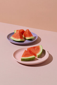 Two plates of freshly cut watermelon on a pink surface with soft shadows and pastel background.