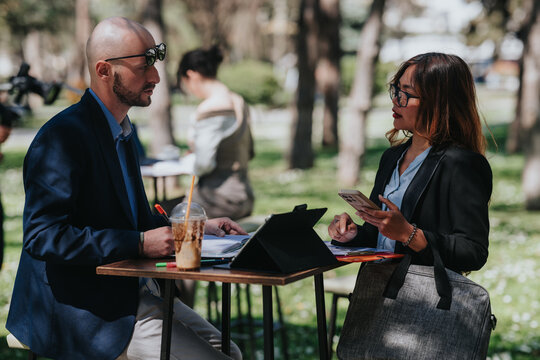 Two business colleagues review documents and digital devices while standing at a small outdoor table. The professional pair consult notes and a smartphone during a casual park meeting.