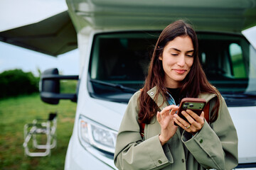 Woman standing outside her recreational vehicle, smiling and texting on her mobile phone, enjoying nature and technology © we.bond.creations