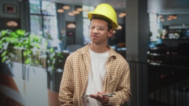 Man handyman wearing yellow hardhat holds wrench with one hand in building interior, plaid shirt and glasses; thoughtful maintenance.