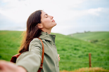 Woman standing in green meadow with eyes closed, breathing deeply and finding peaceful, mindful calm amid rolling hills and fresh air