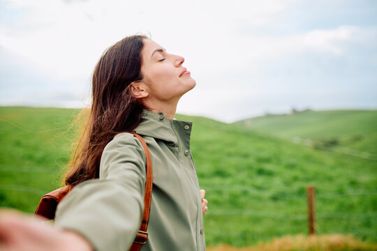 Woman standing in green meadow with eyes closed, breathing deeply and finding peaceful, mindful calm amid rolling hills and fresh air