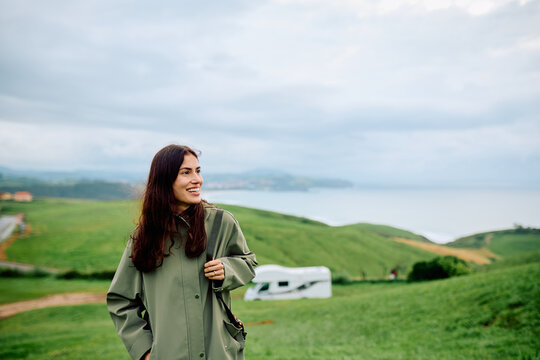 Woman standing on a green hill, smiling and looking at the scenic coast with a camper van in the background