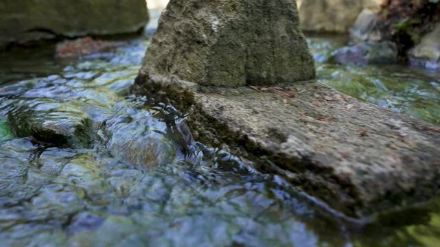 Clear, flowing water gently moves over mossy rocks. Sunlight glints through ripples, creating shimmering reflections. Green algae clings to submerged stones. Scene evokes calmness