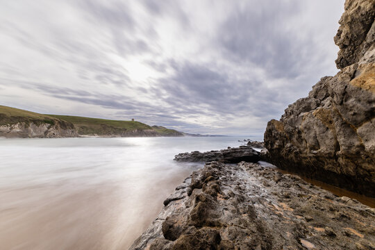 Isolated Tagle Beach at high tide featuring silky water, surrounded by rock formations and green meadows under a cloudy sky with sunbeams. Tagle, Cantabria, Spain.