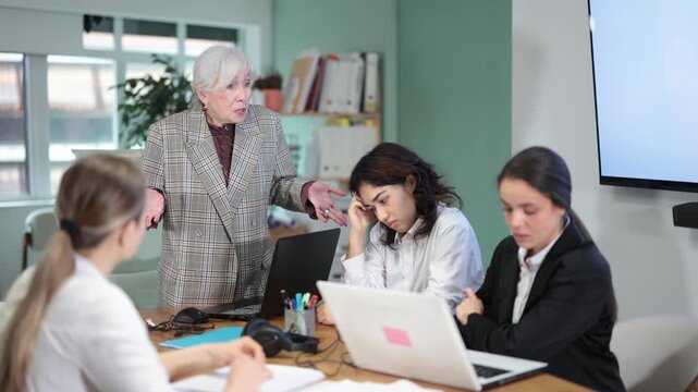  Elderly woman stands at office desk and scolds group of upset women in office