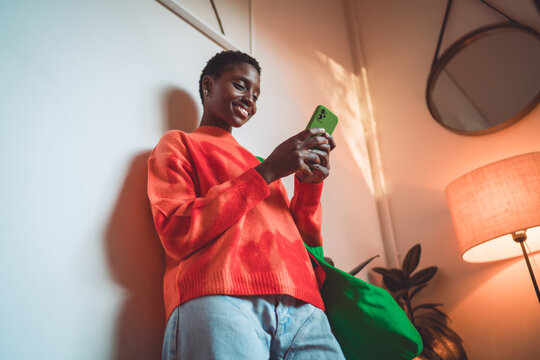 Happy African American woman using mobile app, smartphone 5G connection, orange sweater, green shopper, digital nomad, authentic representation, inclusion, cozy indoor atmosphere, copy space.