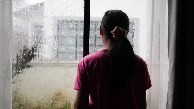 Cinematic back view of a young woman looking out of a window during a rainy day, moody and nostalgic atmosphere with raindrops on glass