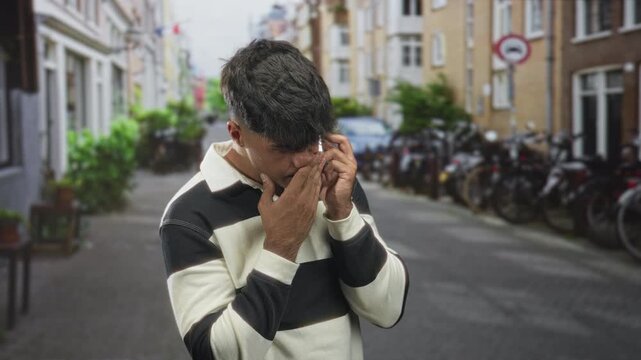 Young hispanic man on street wearing striped sweater holds smartphone to ear and pinches nose with hand; urgent worry.